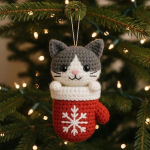 May include: A handmade crochet Christmas ornament shaped like a cat wearing a red mitten. The cat has gray and white fur, a pink nose, and black eyes. The red mitten has a white snowflake design. The ornament hangs from a gold string.