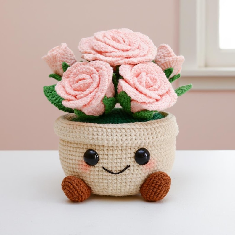 May include: A crocheted flower pot with a smiling face, holding pink roses with green leaves. The pot is beige with brown feet and black eyes. A charming handmade floral decoration.