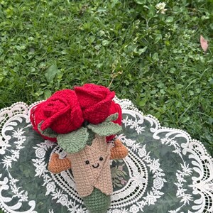 May include: A handmade crochet bouquet featuring two vibrant red roses. The bouquet has green leaves and a smiling, tan-colored stem with small brown accents. It rests on a white lace doily against a backdrop of green grass.