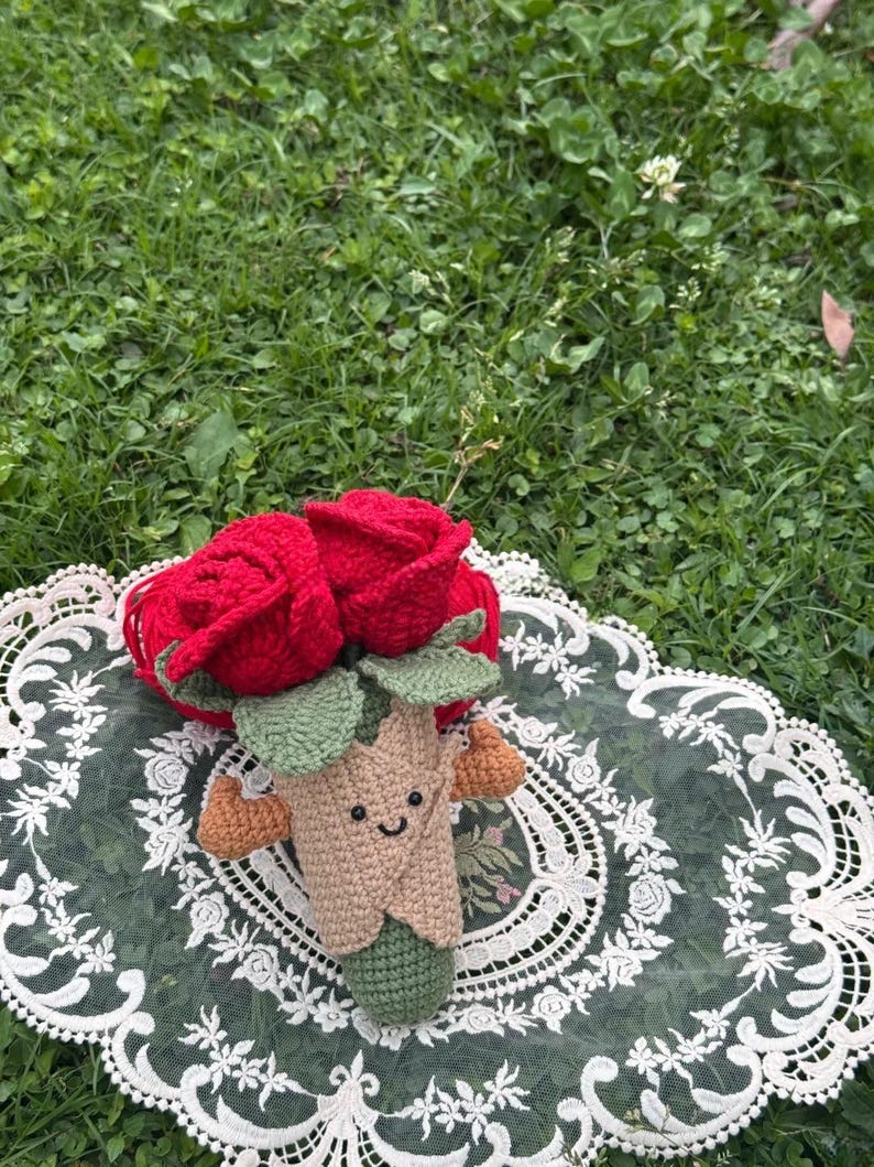 May include: A handmade crochet bouquet featuring two vibrant red roses. The bouquet has green leaves and a smiling, tan-colored stem with small brown accents. It rests on a white lace doily against a backdrop of green grass.