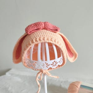 May include: A peach-colored crochet baby bonnet with long bunny ears and a pink bow on top. The bonnet is displayed on a white stand with a decorative lace trim. The ears and bow are a darker shade of pink.