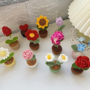 Könnte beinhalten: Eine Sammlung handgefertigter Häkelblumen in kleinen Töpfen. Die Blumen umfassen eine Sonnenblume, eine Tulpe, eine Rose, eine Gänseblümchen und andere, in verschiedenen Farben wie Gelb, Rosa, Rot und Weiß. Die Töpfe sind braun und beige.