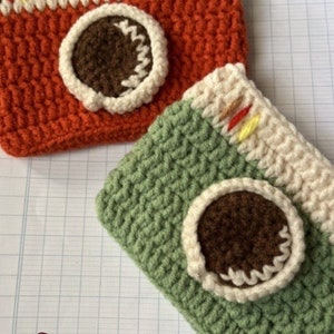 May include: Two crocheted camera-shaped pouches. One is orange, white, and brown. The other is green, white, and brown.
