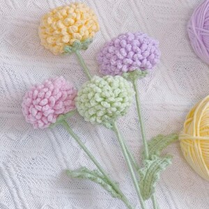 May include: A bouquet of four crocheted flowers in yellow, green, pink, and purple. The flowers are on stems with green leaves and are arranged on a white crocheted surface.