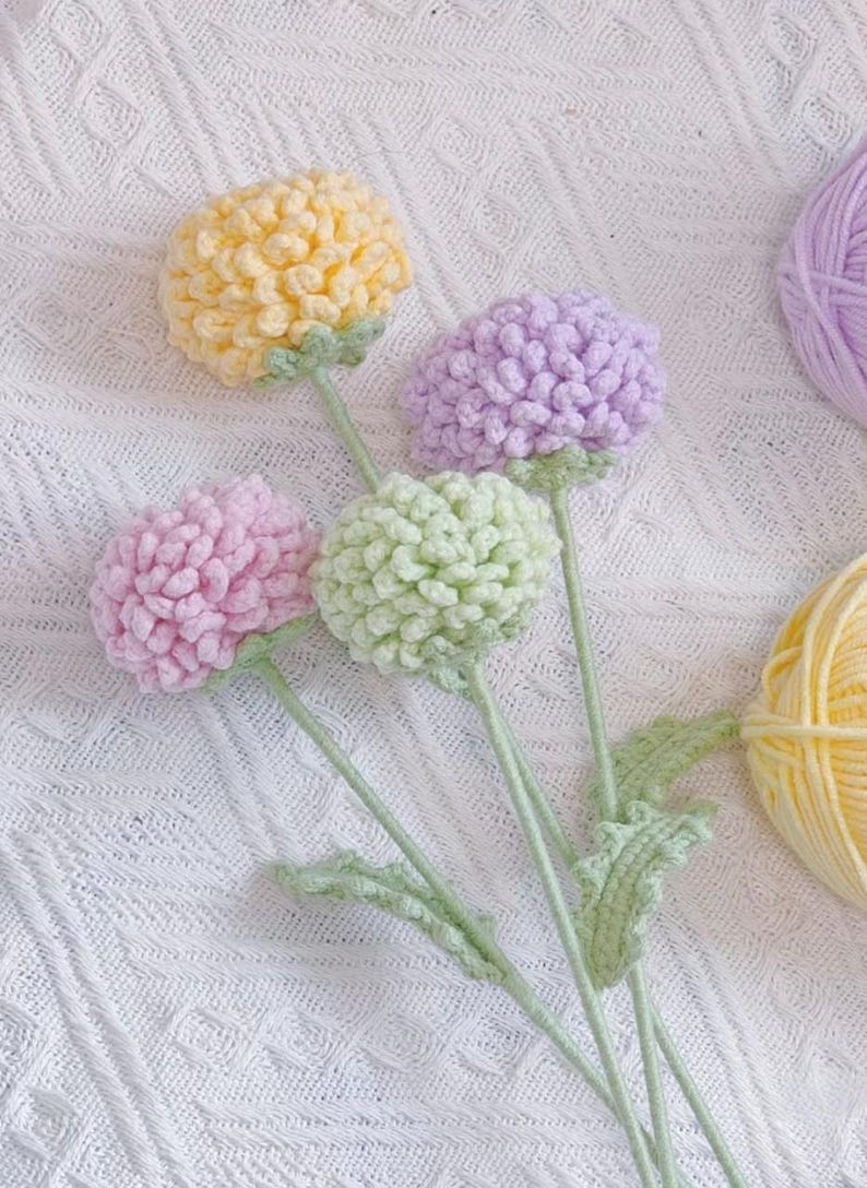 May include: A bouquet of four crocheted flowers in yellow, green, pink, and purple. The flowers are on stems with green leaves and are arranged on a white crocheted surface.