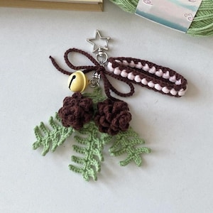 May include: A handmade keychain featuring a star-shaped charm, a small bell, and crocheted elements. The design includes brown pinecones, green fern-like leaves, and a brown and white wrist strap. The keychain is crafted with yarn and metal hardware.