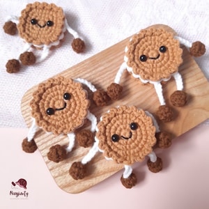 Puede incluir: Cuatro juguetes de crochet con forma de galleta, de color marrón y blanco, con caras sonrientes y piernas marrones y blancas. Los juguetes están dispuestos en una tabla de cortar de madera.