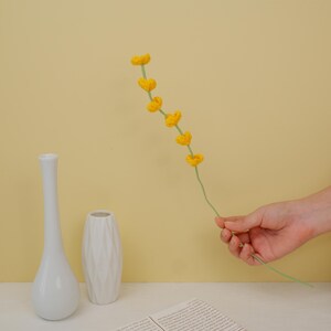 May include: A single stem of yellow crocheted flowers in a hand against a yellow background. Two white ceramic vases are on a table in the foreground.