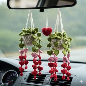 May include: Two crochet hanging planters with green leaves and cascading red and pink heart-shaped accents. A single red heart hangs between the planters. The planters are suspended by white cords, adding a touch of handmade charm.