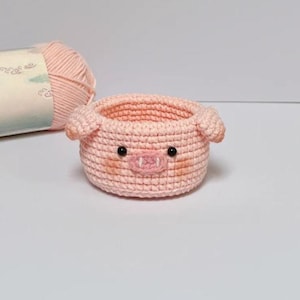 Puede incluir: Una cesta de ganchillo rosa claro hecha a mano con forma de cerdo. La cesta tiene pequeños ojos negros, un hocico rosa y dos pequeñas orejas. Una madeja de hilo rosa está en el fondo. Esta cesta decorativa es ideal para guardar objetos pequeños.