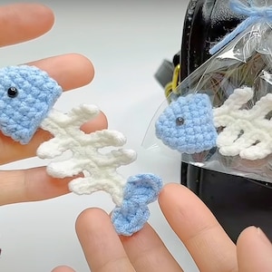 May include: A crocheted blue fish with a white crocheted bone tail. The fish has a black eye and is attached to a silver keychain.