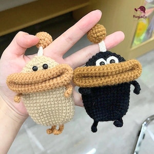 Puede incluir: Dos juguetes de peluche tejidos a crochet con bocas marrones y ojos negros. Un juguete es beige y el otro es negro.