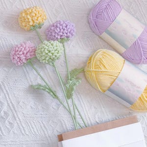May include: A bouquet of crocheted flowers in shades of yellow, green, and pink, with two balls of yarn in purple and yellow. The yarn is labelled in Japanese.