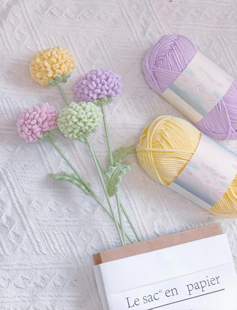 May include: A bouquet of crocheted flowers in shades of yellow, green, and pink, with two balls of yarn in purple and yellow. The yarn is labelled in Japanese.