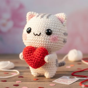 May include: A handmade crochet cat figurine holding a red heart. The cat is white with gray stripes and pink accents on the cheeks and ears. It has black button eyes and a small, smiling mouth. The cat is on a wooden surface.