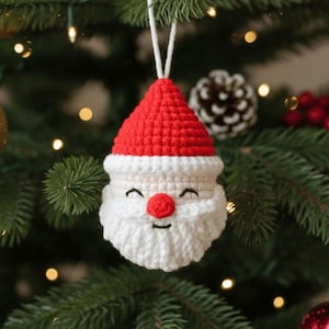 May include: A handmade crochet Christmas ornament shaped like Santa Claus. The ornament features a red hat, a white beard, and a smiling face with a red nose. It is hanging from a white string against a backdrop of a Christmas tree.