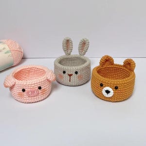 May include: Three handmade crochet baskets in the shape of animals: a pink pig, a gray bunny, and an orange bear. Each basket has a round shape with animal features like ears and a snout. The baskets are on a white surface.