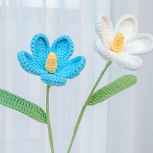 Op de afbeelding: Twee gehaakte bloemen met groene stelen en bladeren. Eén bloem is blauw met een gele kern, de andere is wit met een gele kern.