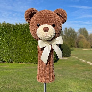 Pode incluir: Uma capa para a cabeça do taco de golfe em forma de urso de peluche castanho com um focinho creme e um laço creme. A capa é feita de um material macio e texturizado. O fundo é um relvado verde e um céu azul.