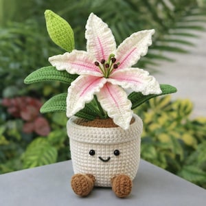 Pode incluir: Um lírio de crochê em um vaso sorridente. A flor tem pétalas brancas com detalhes em rosa e estames marrons. O vaso é branco sujo com pés marrons.