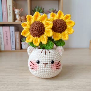 May include: A crocheted cat-shaped planter holding two yellow and brown sunflowers. The white planter has pink cheeks and black eyes. The sunflowers have green leaves and are made of yarn.