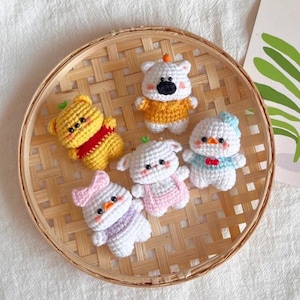 Può includere: Una collezione di sei figurine di animali all'uncinetto fatte a mano, tra cui un orso giallo, un orso bianco, un'anatra, un agnello e un coniglio. Le figurine sono disposte in un cestino intrecciato. Ogni figura ha dettagli unici.