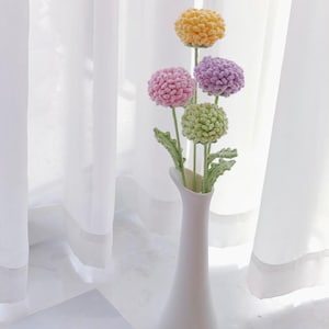 May include: A white vase containing four crocheted flowers in shades of yellow, pink, purple, and green. The flowers are arranged in a bouquet and are sitting on a white surface.