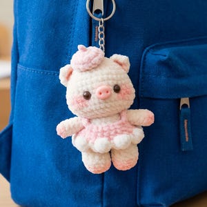 May include: A crocheted pig keychain featuring a cream-colored body with pink accents on the cheeks, ears, and feet. The pig wears a small pink beret and a matching pink tutu skirt, attached to a silver-toned metal keyring and chain.