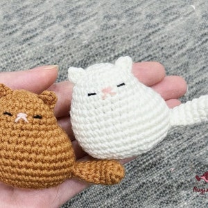 Può includere: Due peluche di gatto all'uncinetto, uno marrone e uno bianco, con occhi neri e nasi rosa. I peluche sono seduti su una mano.