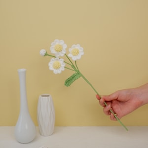 May include: A bouquet of crocheted white daisies with yellow centers and green leaves, held in a hand against a yellow background. Two white ceramic vases are on a white surface.