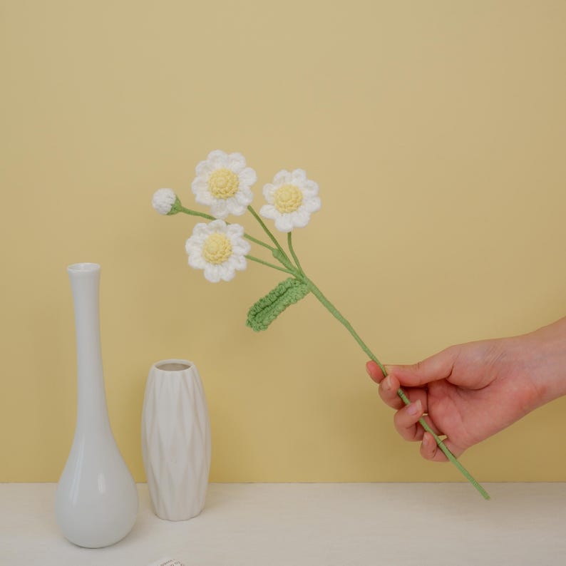 May include: A bouquet of crocheted white daisies with yellow centers and green leaves, held in a hand against a yellow background. Two white ceramic vases are on a white surface.