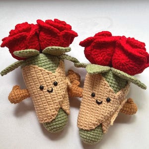 May include: Two handmade crochet rose buds with smiling faces. The red roses sit atop green leaves and tan and green stems. The crochet flowers have small arms and are displayed on a white surface.