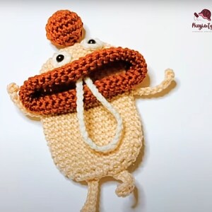 Może przedstawiać: Pluszowa zabawka amigurumi wykonana na szydełku z brązowym kapeluszem, pomarańczowymi ustami i białymi sznurkowymi detalami. Zabawka jest beżowa i ma kapryśny wyraz twarzy.