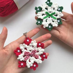 May include: Two handmade crochet snowflakes in white, red, and green. One snowflake has a red and white gingham bow, the other has a green ribbon. A ball of red yarn is in the background. These decorative snowflakes are perfect for holiday decor.