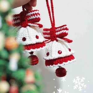Könnte beinhalten: Zwei handgefertigte Weihnachtsglocken-Ornamente aus Häkelarbeit. Jede Glocke ist weiß mit roter Verzierung und einer roten Schleife. Rote Glöckchen hängen von unten. Die Ornamente werden von einer roten Schnur gehalten.