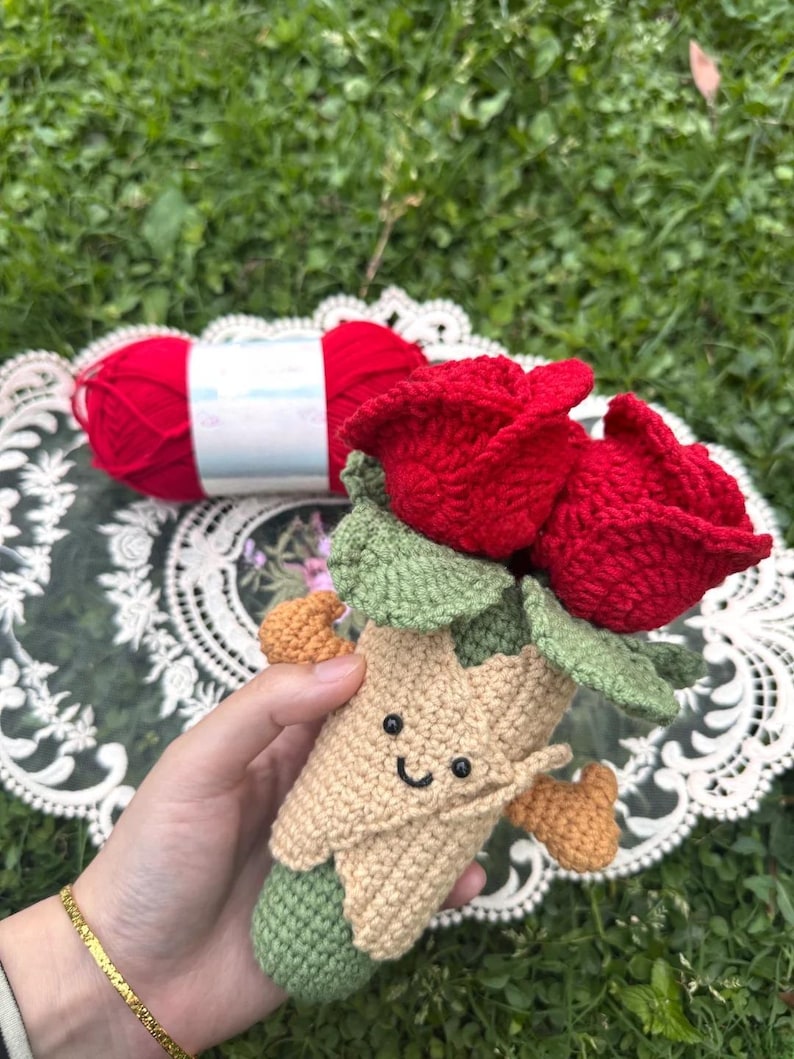 May include: A handmade crochet bouquet featuring two vibrant red roses with green leaves. The bouquet has a tan and green stem with a smiling face. A ball of red yarn is in the background.