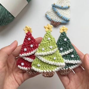 Peut inclure: Ornements de sapin de Noël crochetés en rouge, vert et blanc, chacun avec une étoile jaune et des flocons de neige blancs. Les arbres ont une base marron et sont tenus dans les mains. Une pelote de laine verte est en arrière-plan.