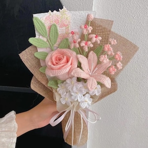 Può includere: Un bouquet all'uncinetto fatto a mano con una grande rosa rosa, un giglio rosa e piccoli fiori bianchi. Il bouquet è avvolto in tela di iuta e legato con un nastro bianco. È inclusa una piccola carta con illustrazioni floreali.