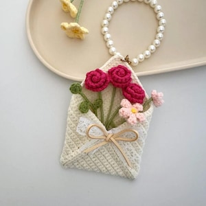 Puede incluir: Una bolsa en forma de sobre de ganchillo color crema llena de flores de ganchillo rosas y rojas. Una pulsera de perlas está unida a la bolsa. Flores amarillas de ganchillo están en el lado izquierdo de la imagen.