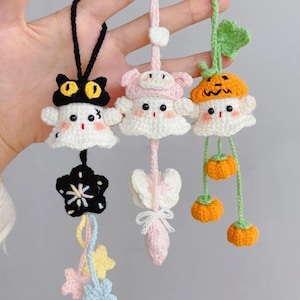 May include: Three handmade crochet ornaments. One is a black cat with yellow eyes, another is a pink character, and the third is a pumpkin. Each has a white body and is suspended by a string. The cat has a black star, the pink character has a bow, and the pumpkin has dangling pumpkins.