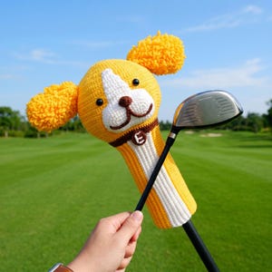 Op de afbeelding: Een gele en witte gehaakte golfclubhoofdbeschermer in de vorm van een hond met flaporen. De hoes heeft een bruine kraag met de letter "E". Een golfclub wordt in de hoes gestoken, vastgehouden door een persoon.