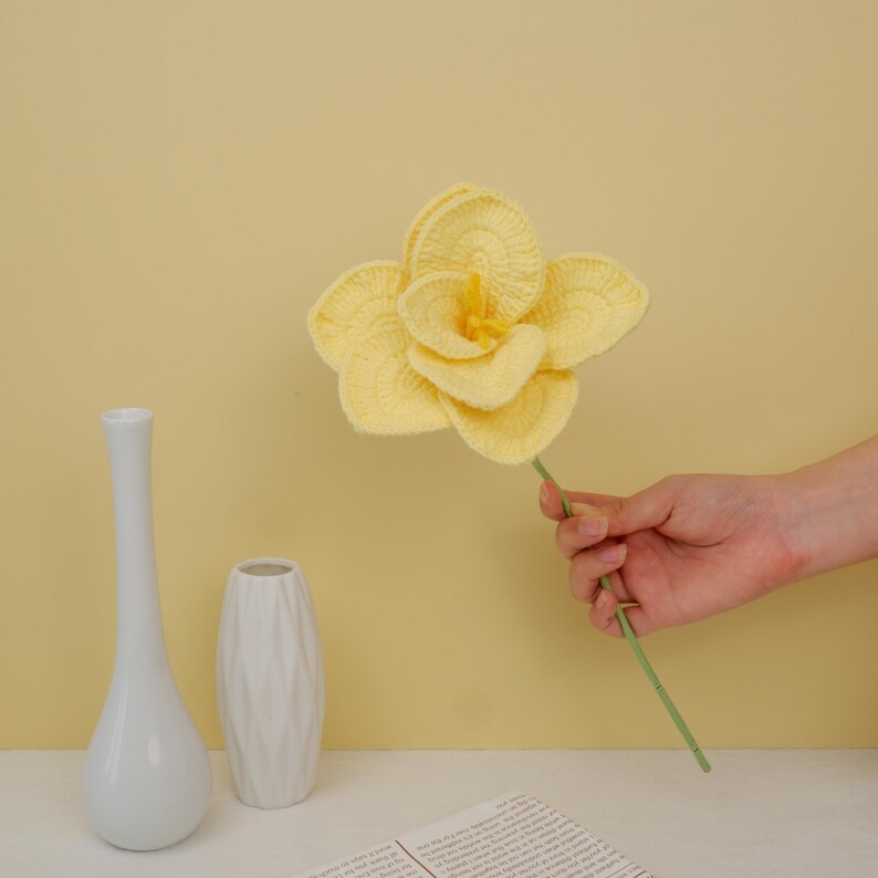 May include: A crocheted yellow flower with a green stem, held in a hand against a yellow background. Two white vases are on a table in the foreground.