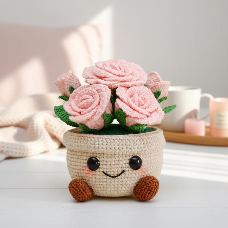 May include: A crocheted flower pot with a smiling face, holding pink crocheted roses with green leaves. The pot is beige with black eyes, a curved mouth, and brown feet. The flowers are arranged in a bouquet.
