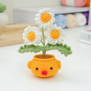 Puede incluir: Un arreglo floral de margaritas de ganchillo en una maceta amarilla con forma de pollito. Las margaritas tienen pétalos blancos y centros amarillos. La maceta tiene pequeños ojos negros y un pico rojo. Las hojas y los tallos verdes añaden un toque natural.