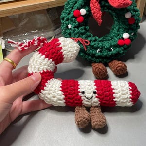 May include: Handmade Christmas decorations. A crocheted candy cane in red and white stripes with a smiling face and brown feet, and a green wreath with red accents and brown feet.