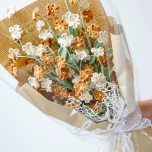May include: A bouquet of crocheted flowers in shades of white, brown, and green, wrapped in brown paper and white lace. The flowers are arranged in a vase and are tied with a white ribbon. The bouquet is sitting on a white surface.