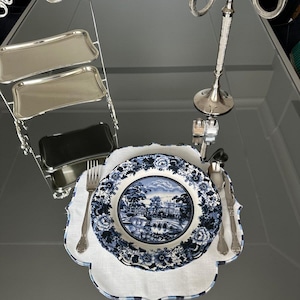 Pode incluir: Uma mesa formal com um prato com estampa floral azul e branco com uma cena de paisagem, talheres e um sousplat com bordas onduladas. Uma bandeja de servir prateada em camadas e um castiçal também estão presentes, criando um arranjo de jantar elegante.