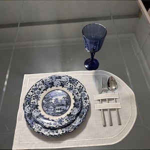 Pode incluir: Uma mesa posta com pratos florais azuis e brancos, um copo de vinho azul e talheres sobre um sousplat branco. Os pratos têm uma cena de paisagem no centro.