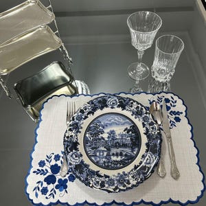 Pode incluir: Uma mesa formal com um prato com padrão floral azul e branco, um tapete branco com bordado floral azul e talheres. Também estão presentes dois copos de cristal e uma bandeja de servir prateada.