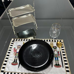 Op de afbeelding: Een tafelsetting met een zwart bord, goudkleurig bestek en een decoratieve placemat met een notenkrakerontwerp. Een etagère en een wijnglas met een gouden rand zijn ook aanwezig.